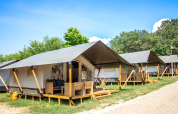 Rij moderne Safari Tent Comfort met veranda’s, omgeven door bomen en gras onder een blauwe lucht.