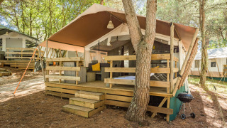 Luxury safari tent with wooden deck, outdoor seating, and grill set in a tranquil, forested campsite.