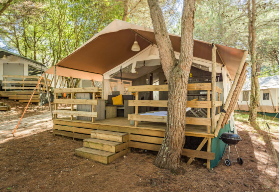 Luxury safari tent with wooden deck, outdoor seating, and grill set in a tranquil, forested campsite.