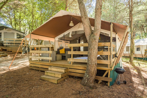 Tente safari de luxe avec terrasse en bois, coin salon extérieur et barbecue dans un cadre boisé paisible.