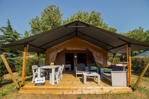 Safari tent Comfort met airconditioning, houten veranda, tuinmeubilair, tafel en stoelen in groene omgeving.