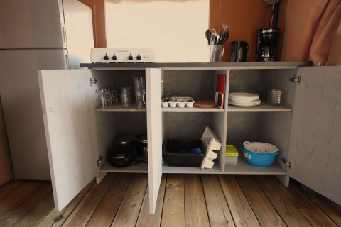 Credenza da cucina aperta con stoviglie e utensili in una tenda safari Comfort con aria condizionata al Camping Porto Sole.