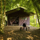 Tienda tipo safari en el bosque, con un kart de pedales delante y rodeada de árboles verdes y frondosos.