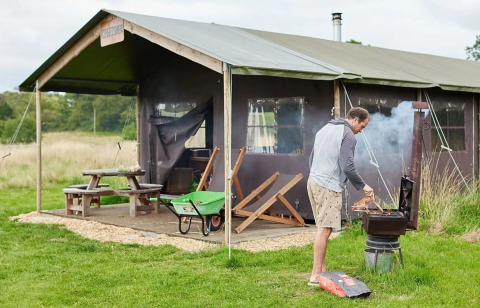 Homme faisant un barbecue devant une tente safari à Feather Down Heydon Grove Farm, UK, table en fond.