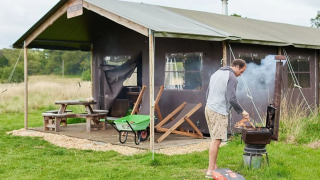 Man barbecue voor safaritent bij Feather Down Heydon Grove Farm, VK, picknicktafel op de achtergrond.