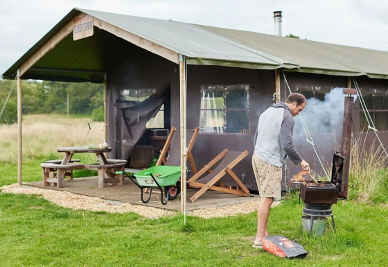 Homme faisant un barbecue devant une tente safari à Feather Down Heydon Grove Farm, UK, table en fond.