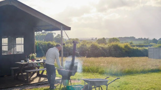 Mann grillt neben einer Safarizelt-Hütte auf einer sonnigen Wiese mit Blick auf Felder und Bäume.