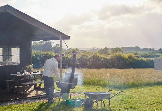 Mann grillt neben einer Safarizelt-Hütte auf einer sonnigen Wiese mit Blick auf Felder und Bäume.