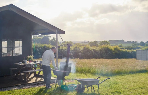 Homme faisant un barbecue près d'une cabane-tente safari par temps ensoleillé, champs et arbres au fond.