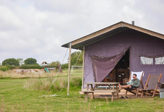 Hombre relajándose fuera de tienda safari con ducha privada en Feather Down New Barn Farm, Reino Unido.
