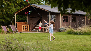 Bambini fanno volare un aquilone sul prato di fronte a una tenda safari immersa nella natura e alberi.