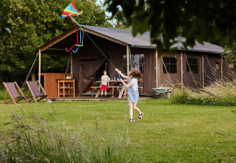 Bambini fanno volare un aquilone sul prato di fronte a una tenda safari immersa nella natura e alberi.