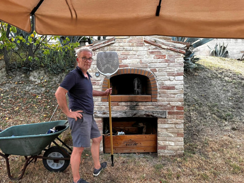 Hombre con pala para pizza frente a un horno de ladrillo al aire libre en Agriturismo Angeli Sognanti, Marche, Italia.
