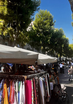 Mercado al aire libre con puestos de ropa y gente caminando bajo árboles en Marche, Italia, cerca de Angeli Sognanti.