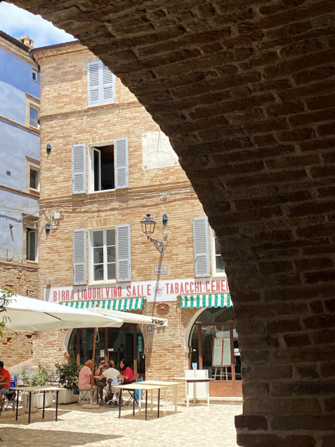 Vista attraverso un arco in pietra su una piazza con caffè a Cossignano, Marche, Italia, in una giornata soleggiata.