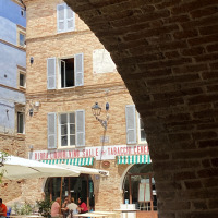 Vista a través de un arco de piedra hacia una plaza y cafetería en Cossignano, Marche, Italia, en día soleado.