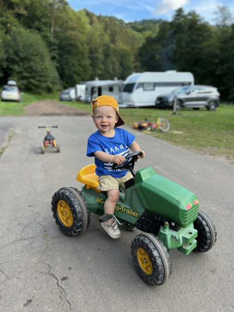 Un niño pequeño monta un tractor de juguete verde en Camp Kyllburg, con caravanas y vegetación alrededor.