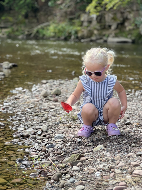 Bambina con occhiali da sole gioca sulla riva sassosa del fiume al Camp Kyllburg, Renania-Palatinato.
