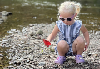 Kleines Mädchen mit Sonnenbrille spielt am steinigen Flussufer im Ferienpark Camp Kyllburg, Rheinland-Pfalz.