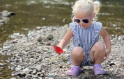 Young girl in sunglasses and a striped outfit playing with a red object by a rocky river at Camp Kyllburg.