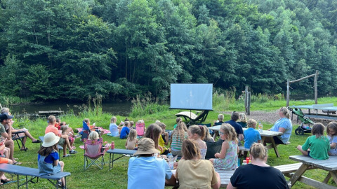 Bambini e adulti guardano un film all'aperto al Camp Kyllburg, vicino a un fiume tra alberi verdi.