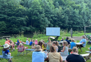 Børn og voksne ser udendørs film på Camp Kyllburg, omgivet af grønt skovområde ved en flod.