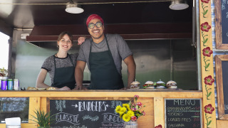 Dos vendedores sonrientes ofrecen hamburguesas en un food truck decorado con flores en un parque vacacional.