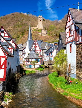 Casas de entramado de madera junto a un arroyo en Kyllburg, Alemania, con colinas y un castillo en ruinas.