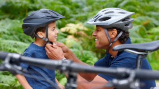 Un adulto ayuda a un niño a ponerse el casco de bicicleta al aire libre, rodeados de bicicletas y vegetación.