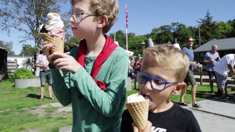 Dos niños comen helados en conos en Rosenvold Strand Camping, Dinamarca, rodeados de gente en un día soleado.