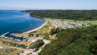 Luftfoto af Rosenvold Strand Camping i Danmark, med campingvogne, skov, strand og småbådehavn.