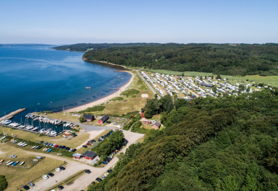 Luftfoto af Rosenvold Strand Camping i Danmark, med campingvogne, skov, strand og småbådehavn.