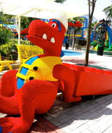 Statue de dinosaure colorée en briques LEGO dans un parc avec des bancs jaunes et des palmiers autour.