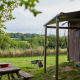 Udsigt fra et safaritelts terrasse mod grønne marker, træer og picnicbord på en rolig sommerdag.