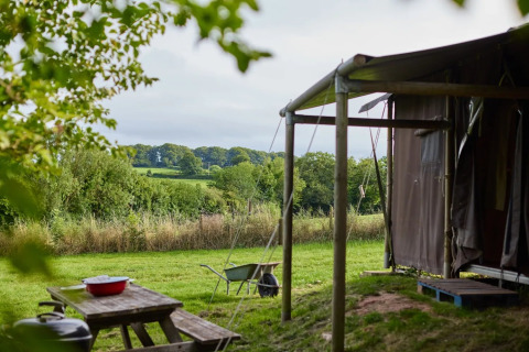 Uitzicht vanuit een safaritent op groene velden, bomen en een picknicktafel tijdens een zomerse dag.