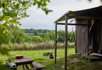 Ausblick von einem Safarizelt auf grüne Felder, Bäume und Picknicktisch an einem ruhigen Sommertag.