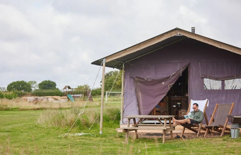 Man zit lezend buiten een open safaritent op gras met picknicktafel onder een bewolkte lucht.