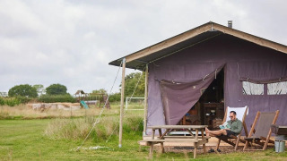 Hombre sentado leyendo frente a una tienda de safari abierta en un campo con mesa de picnic y césped.