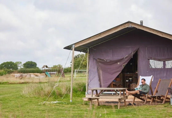 Hombre sentado leyendo frente a una tienda de safari abierta en un campo con mesa de picnic y césped.