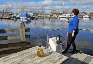 Mujer en una casa flotante en Marina Heeg, Países Bajos, sosteniendo una copa de vino y mirando el puerto.