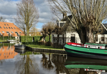 Photo d’un hébergement glamping au bord d’un canal, avec bateaux, arbres dénudés et toit rouge brique.