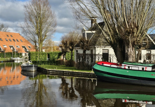 Foto van glamping-accommodatie aan een kanaal met boten, rood dak en kale bomen in de winter.