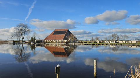 Casa flotante en Marina Heeg, Países Bajos, reflejada en el agua con nubes, árboles y cielo azul.