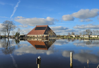 Houseboat at Marina Heeg, Netherlands, reflected in calm water with clouds, trees, and blue sky.