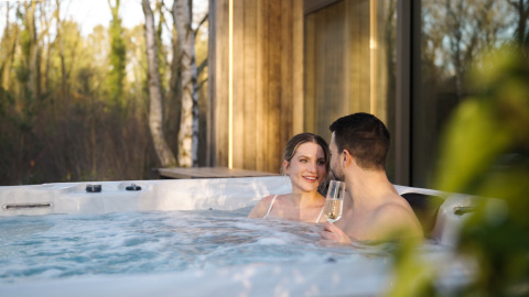 Couple relaxing in a jacuzzi at Lodge with Jacuzzi, toasting with champagne and surrounded by trees.