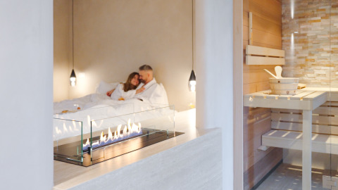Couple in white robes relaxing by a fireplace in the Wellness Suite at Woodz Lodges with sauna in Belgium.