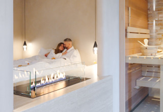 Pareja en batas blancas relajándose junto a la chimenea en la Wellness Suite de Woodz Lodges con sauna en Bélgica.