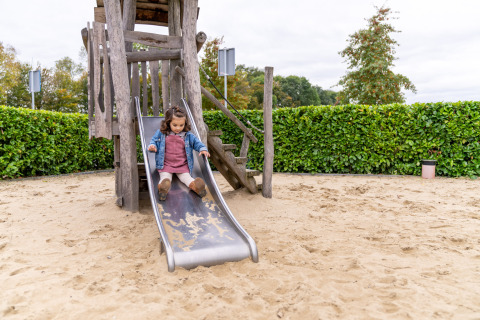 Kleines Mädchen rutscht eine Holzrutsche auf einem Spielplatz mit Sandboden bei einer Lodge entlang.