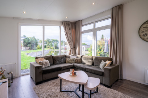Moderne lodge-stue med stor hjørnesofa, naturudsigt gennem vinduer, lyse gardiner og sofabord.