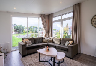 Moderne lodge-stue med stor hjørnesofa, naturudsigt gennem vinduer, lyse gardiner og sofabord.